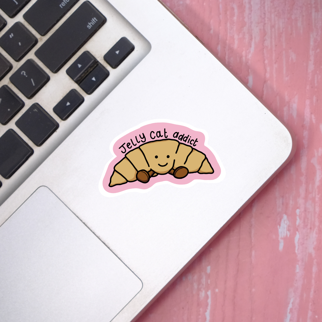 Sticker on a laptop keyboard with a croissant design and 'Jelly cat addict' text on a pink surface.