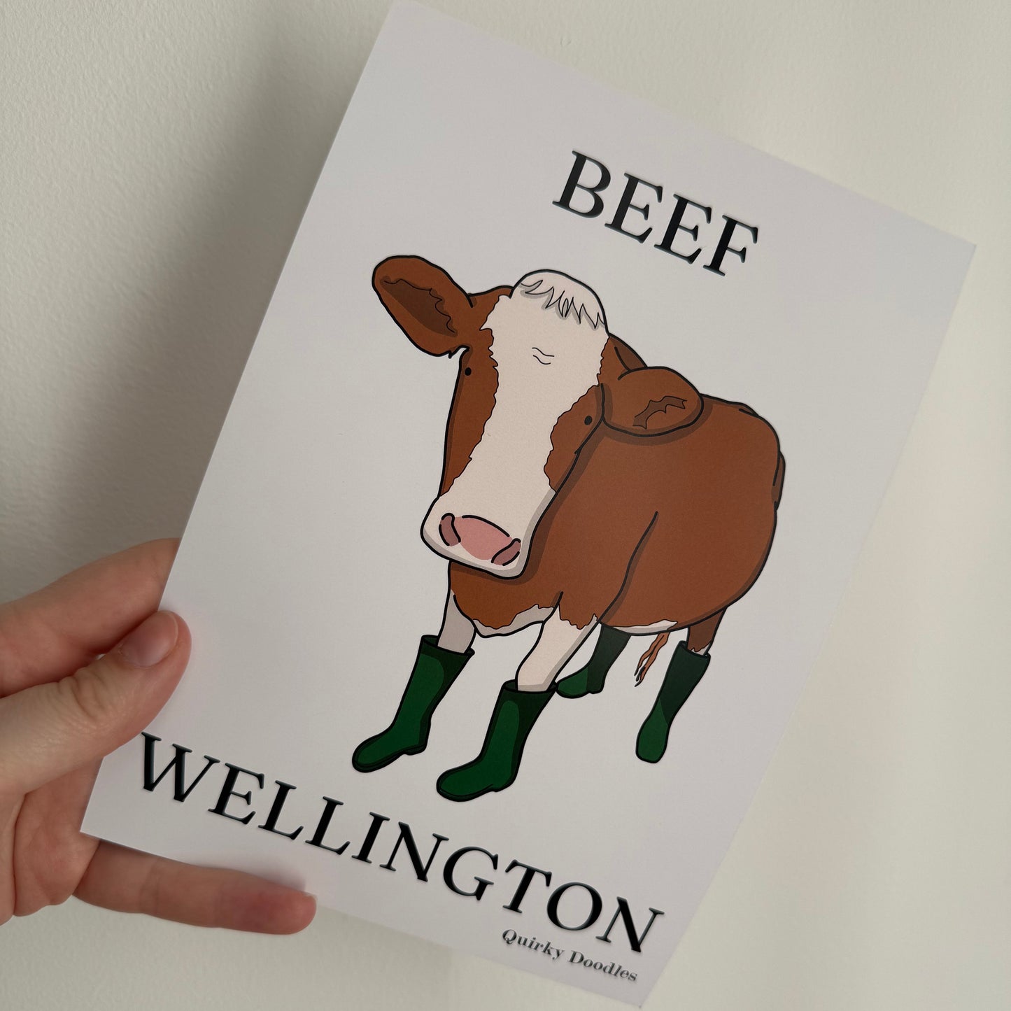Print with a cartoon cow and text 'Beef Wellington' held by a hand on a light background