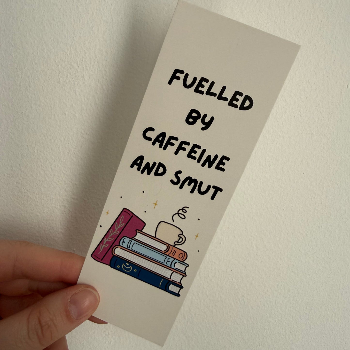 Bookmark with text 'Fueled by caffeine and smut' held against a plain background