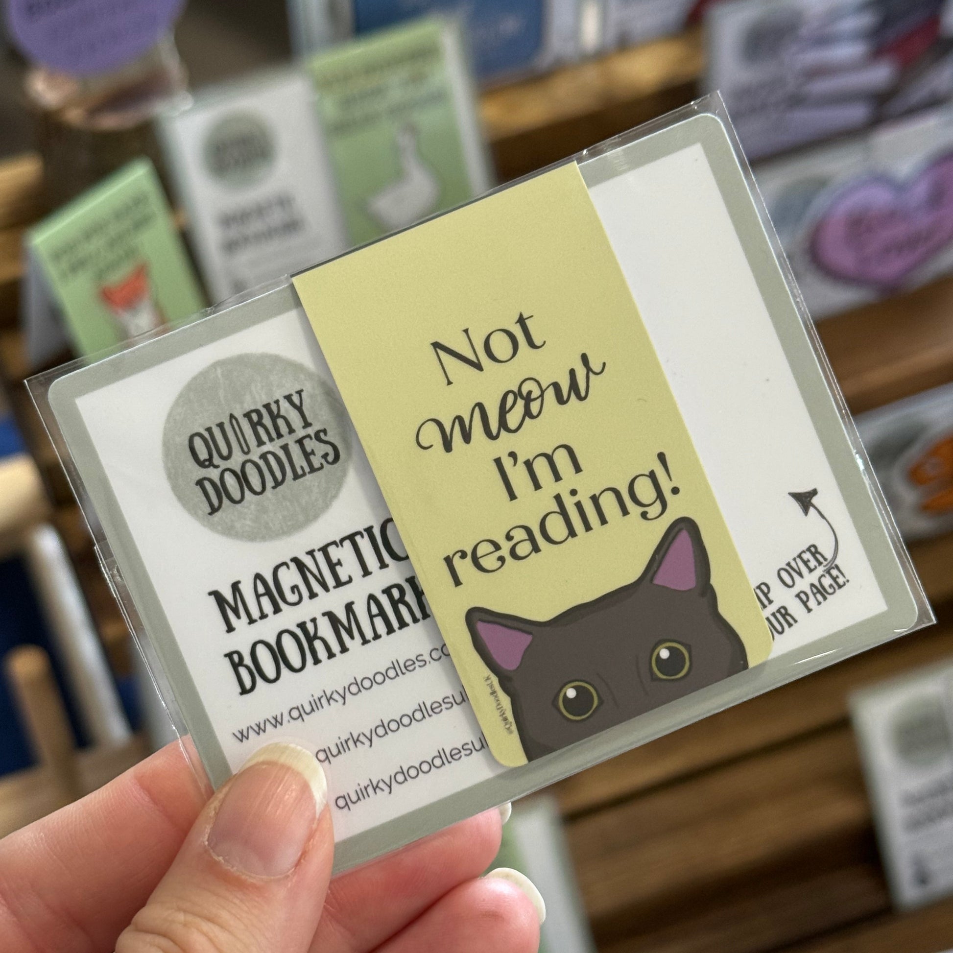 Hand holding a magnetic bookmark with a cat illustration and text, with a blurred background of similar products.