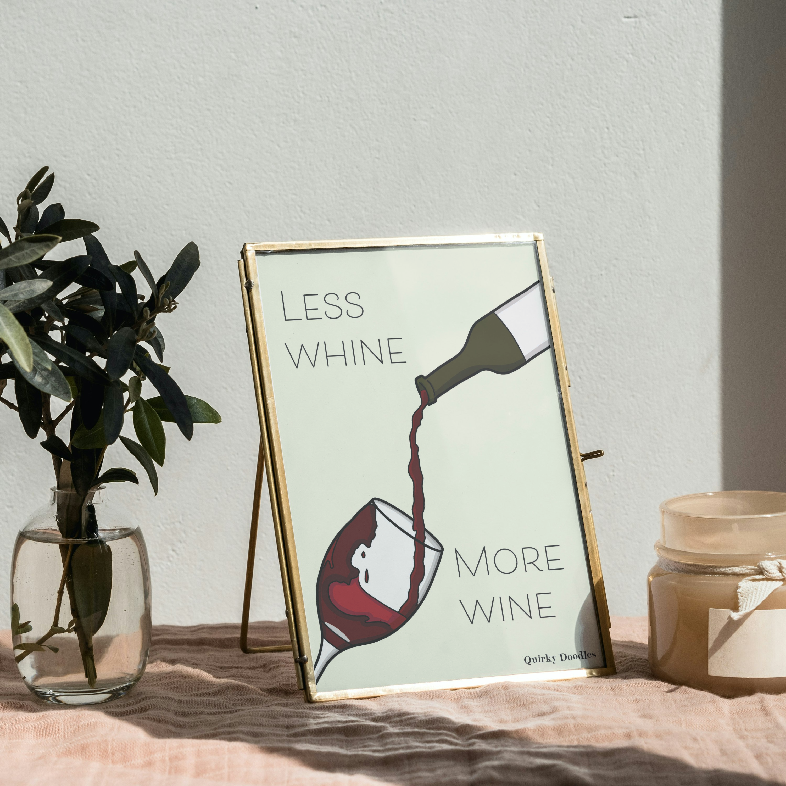 Framed print with 'Less Whine, More Wine' design on a table with a plant and candle.
